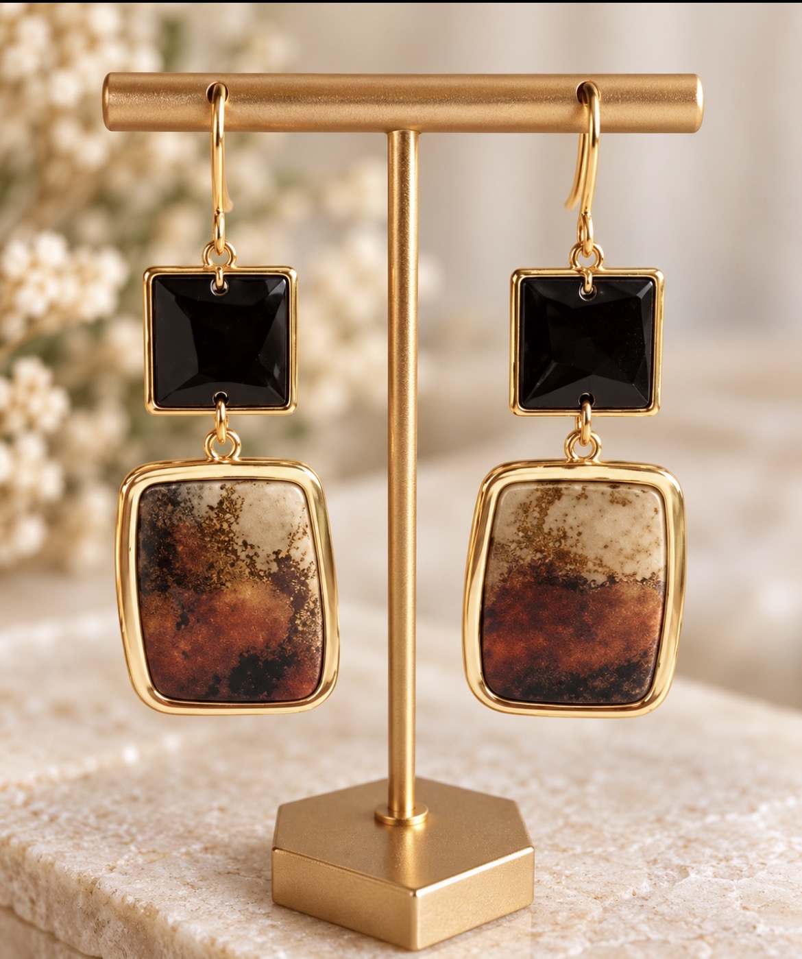Gold-rimmed square drop earrings with black gemstone tops and marbled cream-and-rust clay, on a gold stand
